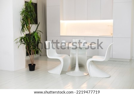 Stylish white dining room with large round table and designer chairs, cups coffee, large houseplant in pot. White plastic chairs pantone. Modern Kitchen interior design with furniture. 
