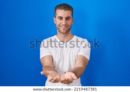 Young caucasian man standing over blue background smiling with hands palms together receiving or giving gesture. hold and protection 