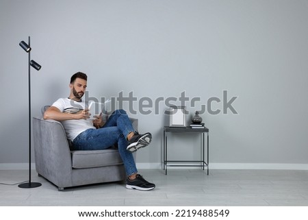 Man reading book in armchair near gray wall, space for text