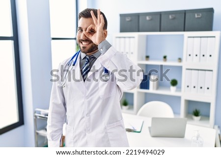 Handsome hispanic man wearing doctor uniform and stethoscope at medical clinic smiling happy doing ok sign with hand on eye looking through fingers 