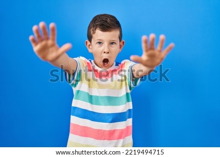 Young caucasian kid standing over blue background doing stop gesture with hands palms, angry and frustration expression 