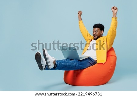 Full body smiling happy young man of African American ethnicity wear yellow shirt sit in bag chair hold use work on laptop pc computer do winner gesture isolated on plain pastel light blue background