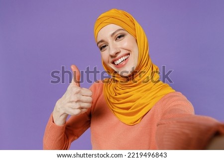 Close up young arabian asian muslim woman in abaya hijab yellow clothes do selfie shot pov on mobile phone show thumb up like gesture isolated on plain pastel light violet background studio portrait