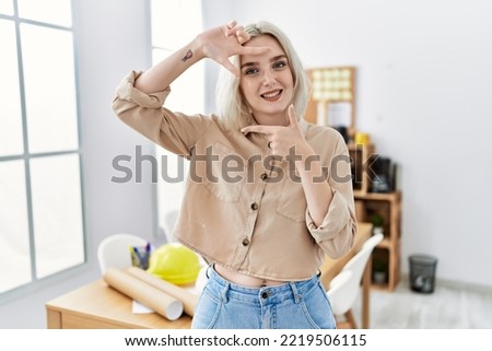 Young beautiful caucasian woman at construction office smiling making frame with hands and fingers with happy face. creativity and photography concept. 