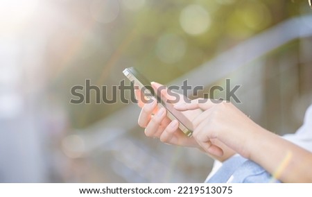 Crop shot of a woman's hand holding a smartphone against a natural green background in evening light. A woman reading a message on a mobile phone during the city