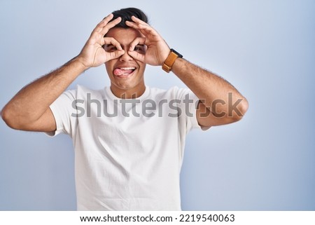 Hispanic man standing over blue background doing ok gesture like binoculars sticking tongue out, eyes looking through fingers. crazy expression. 