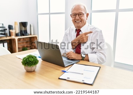 Senior man working at the office using computer laptop cheerful with a smile on face pointing with hand and finger up to the side with happy and natural expression 