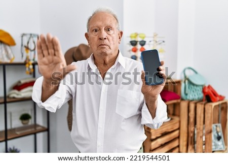 Senior man holding smartphone at retail shop with open hand doing stop sign with serious and confident expression, defense gesture 