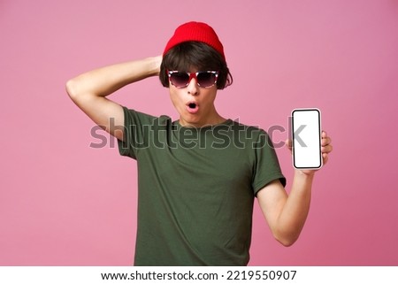 Excited teenager guy hold smartphone with white mock up screen wearing green khaki t-shirt and red hat in Great Britain flag sunglasses isolated on pink background. Mobile app advertisement mock up. 