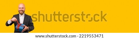 Banner size image of cheerful smiling bearded man in suit opening grift box over yellow background