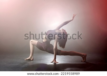 Flexible young female woman doing excersice in fog smoke. Girl wearing fitness dance clothing making dance element performance on isolated black background with red light. Stretching
