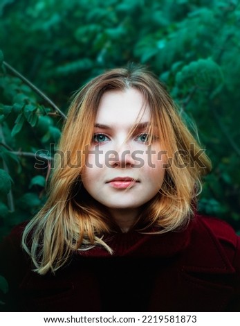 A close-up portrait of a beautiful young blonde woman in a burgundy coat against the background of green foliage of trees. Lifestyle