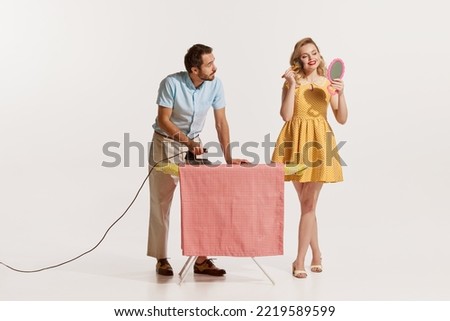 Portrait of man doing ironing while beautiful woman doing her makeup isolated over white background. Concept of retro style, domestic duties, old-fashion, lifestyle. Copy space for ad
