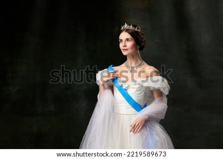 Historical reconstruction. Young queen. Portrait of adorable girl in image of medieval royal person in renaissance style dress isolated on dark background. Comparison of eras, beauty, history, art. Royalty-Free Stock Photo #2219589673