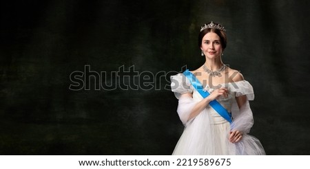 Historical reconstruction. Young queen. Portrait of adorable girl in image of medieval royal person in renaissance style dress isolated on dark background. Comparison of eras, beauty, history, art. Royalty-Free Stock Photo #2219589675