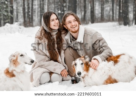 Love romantic young couple girl, guy in snowy cold winter forest walking with pet, dog of hunting breed russian borzoi. Sighthound, wolfhound owner. Having fun, laughing. Stylish fur coat, woolen hat. Royalty-Free Stock Photo #2219601011