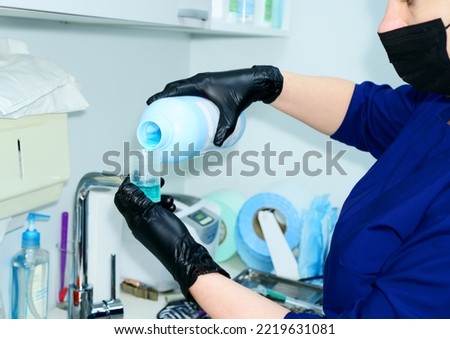 The dentist pours the disinfectant solution from a bottle into a small container. A health care worker wearing black protective gloves prepares to disinfect          