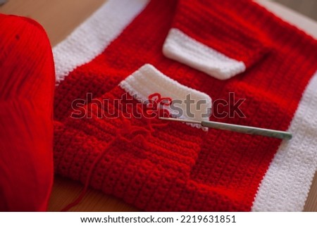 Crocheting red and white Christmas sweater outfit for a baby as Xmas gift watching online tutorial on laptop. Holiday season gifts preparation. DIY hobby making handmade clothes.