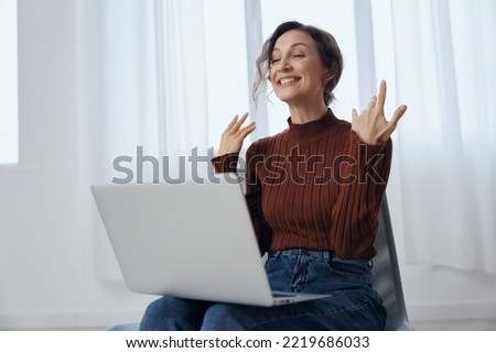 Excited cheerful happy enthusiastic shocked curly awesome young woman talks with friend in video conference call tells about latest cool news holds laptop raise hand active gesturing in chair at home