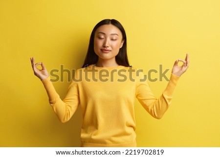 Peaceful Woman Meditating at Studio. Portrait of Asian Young Girl Relaxing and Doing Meditation Gesture with Fingers, Practicing Yoga. Indoor Studio Shot Isolated on Yellow Background