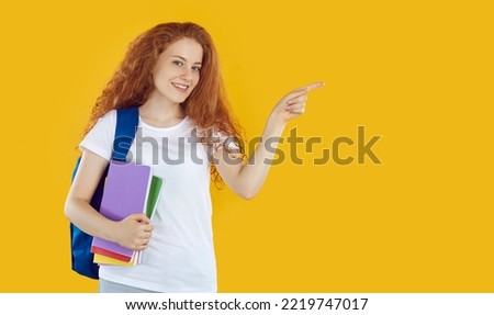 Beautiful female student poiting with her index finger to right side, isolated on orange background. Cute girl with red wavy hair holds notebooks and backpack and points to place for text. Web banner.
