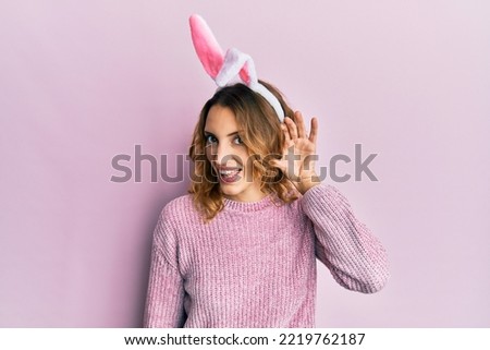 Young caucasian woman wearing cute easter bunny ears smiling with hand over ear listening and hearing to rumor or gossip. deafness concept. 