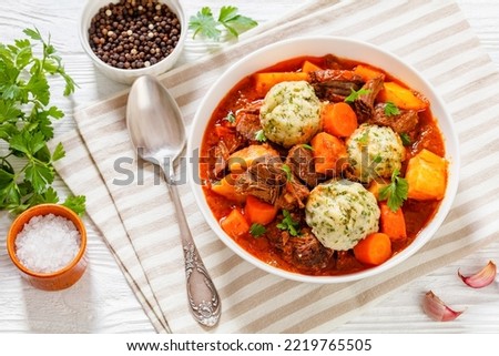 Beef Stew with Dumplings and vegetables in rich tomato and stock based gravy in white bowl on wood table, horizontal view from above, flat lay, free space
