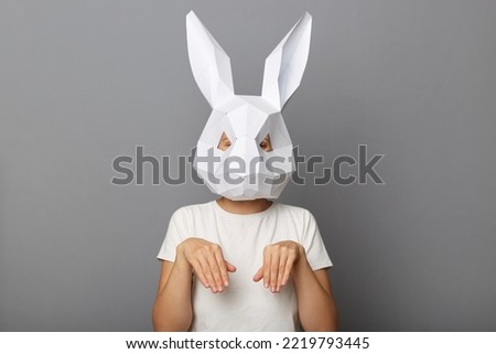Portrait of funny unknown female wearing bunny mask and white t-shirt standing with hare paws, expressing festive emotions, isolated over gray background.