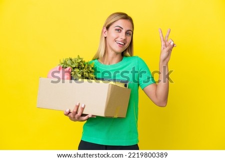 Young Uruguayan girl making a move while picking up a box full of things isolated on yellow background smiling and showing victory sign