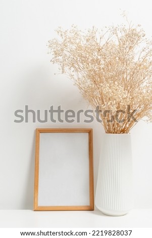 Wooden vertical frame with white vase of gypsophila flowers over white wall. Mockup Template for your design, text.
