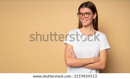 Student girl in glasses and white mockup t-shirt standing with folded arms on brown studio background looking with smile on copy space for your text on the right giving tips for college applicants