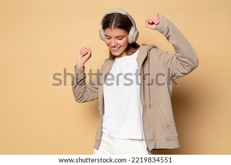 Pretty woman having fun dancing in wireless headphones, hoodie and mockup t-shirt, listening ti pop music or hip-hop rhythms on light brown background