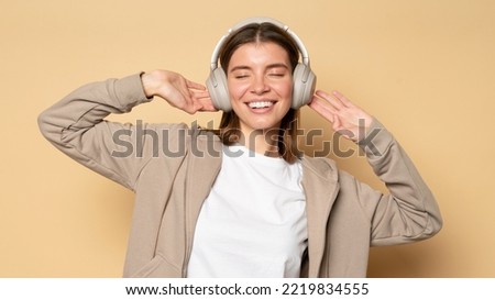 Studio portrait of happy relaxed woman with closed eyes wearing wireless headphones posing on light brown background enjoying popular music