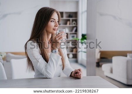 Confident blonde businesswoman sitting at kitchen table at hole drinks water looks aside against blurry interior. Successful Italian fit woman satisfied by healthy lifestyle. Business and healthcare. Royalty-Free Stock Photo #2219838267