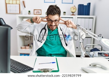 Young man with beard wearing doctor uniform and stethoscope at the clinic doing peace symbol with fingers over face, smiling cheerful showing victory 