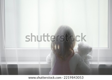 Little girl holding white teddy bear standing alone at window behind transparent day curtains and looking out from home. Back view. Waiting concept. Close up. Royalty-Free Stock Photo #2219843053