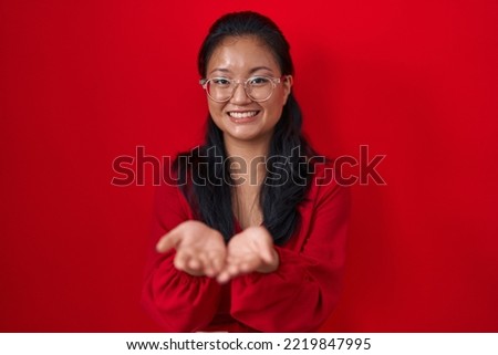Asian young woman standing over red background smiling with hands palms together receiving or giving gesture. hold and protection 