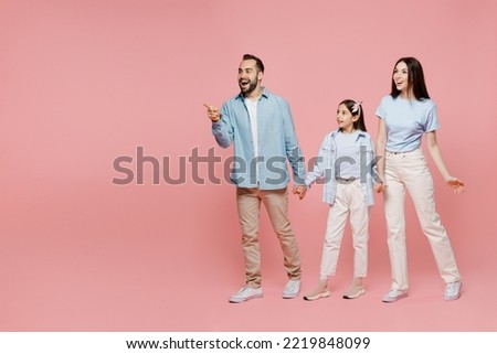 Full body young happy parents mom dad with child kid daughter teen girl in blue clothes hold hands walk going point index finger aside on workspace area isolated on plain pastel light pink background.