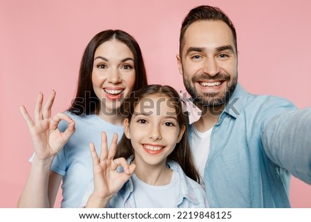 Close up young happy fun smiling parents mom dad with child kid daughter teen girl in blue clothes doing selfie shot pov on mobile cell phone show okay isolated on plain pastel light pink background.
