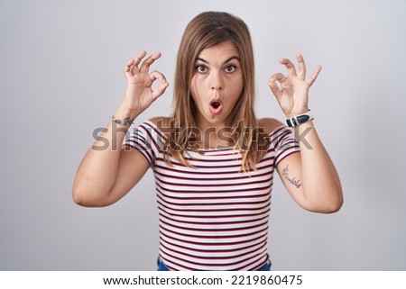 Young hispanic woman standing over isolated background looking surprised and shocked doing ok approval symbol with fingers. crazy expression 