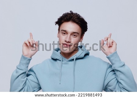 horizontal portrait of a pleasantly smiling young man in a light hoodie pointing his fingers towards an empty space on the background