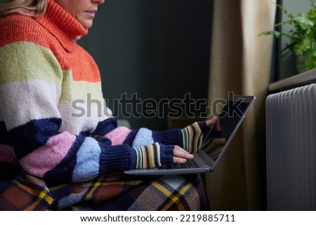 Woman In Gloves With Laptop Trying To Keep Warm By Radiator During Cost Of Living Energy Crisis Royalty-Free Stock Photo #2219885711