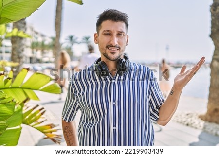Young handsome man listening to music using headphones outdoors smiling cheerful presenting and pointing with palm of hand looking at the camera. 
