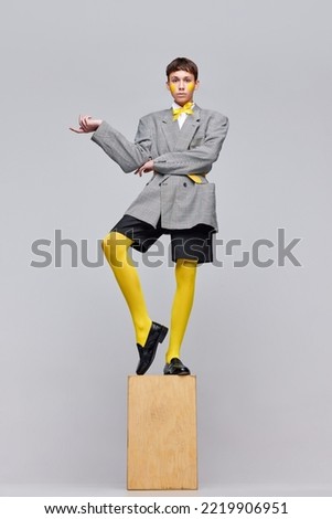 Portrait of stylish boy posing in jacket, blazer and yellow tights isolated over grey background. Yellow blushes . Concept of modern fashion, art photography, style, queer, uniqueness, ad