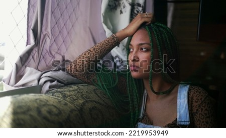 One thoughtful black latina sitting at home couch looking out window with pensive expression