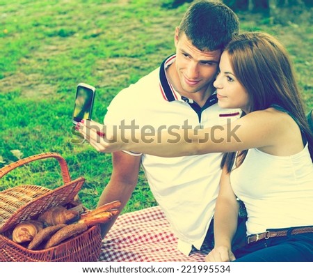 Young couple sitting in the park on a summer day. Picnic basket in the background. Taking picture of themselves with the phone..