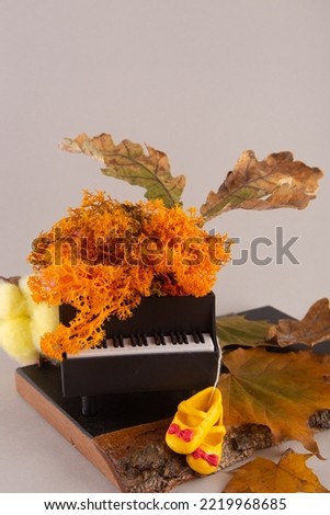 Autumn composition. Small toy black piano and fall decorations. Orange color moss, yellow cotton flower, dry oak leaves and tender dancing shoes on grey background. Vertical photo.