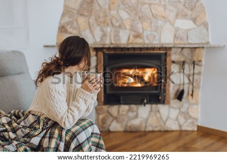 Young woman sitting at home by the fireplace with a hot tea or coffee mug and warming her hands, she is wearing white woollen sweater. Cold houses in Europe concept during energy and gas crisis. Royalty-Free Stock Photo #2219969265