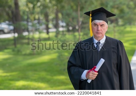 Serious old man in graduation gown holding diploma outdoors.