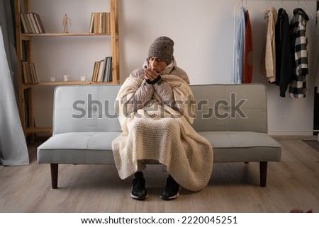 Unwell man in hat and blanket sit in cold living room suffer from heat lack. Unhealthy young man struggle from chill freeze at home. No heating concept.